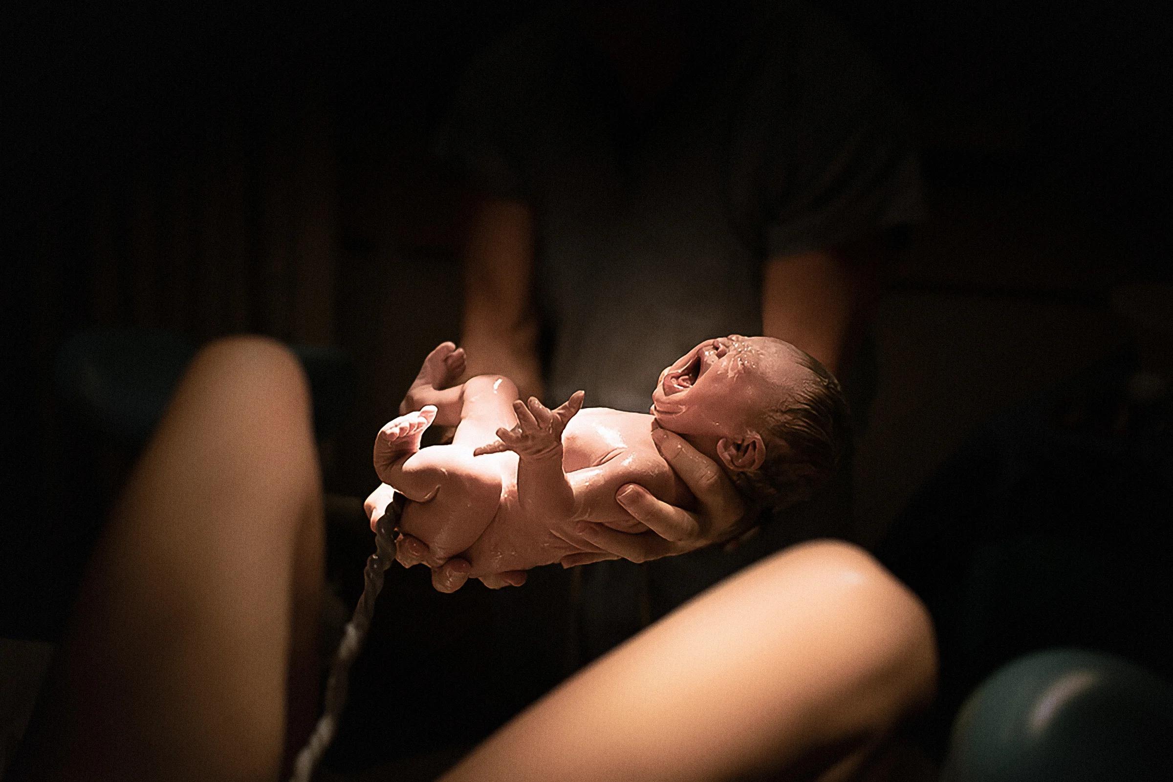 A newborn, moments old, held up in a pair of hands in a dim birth room, the mother’s knees in the foreground.